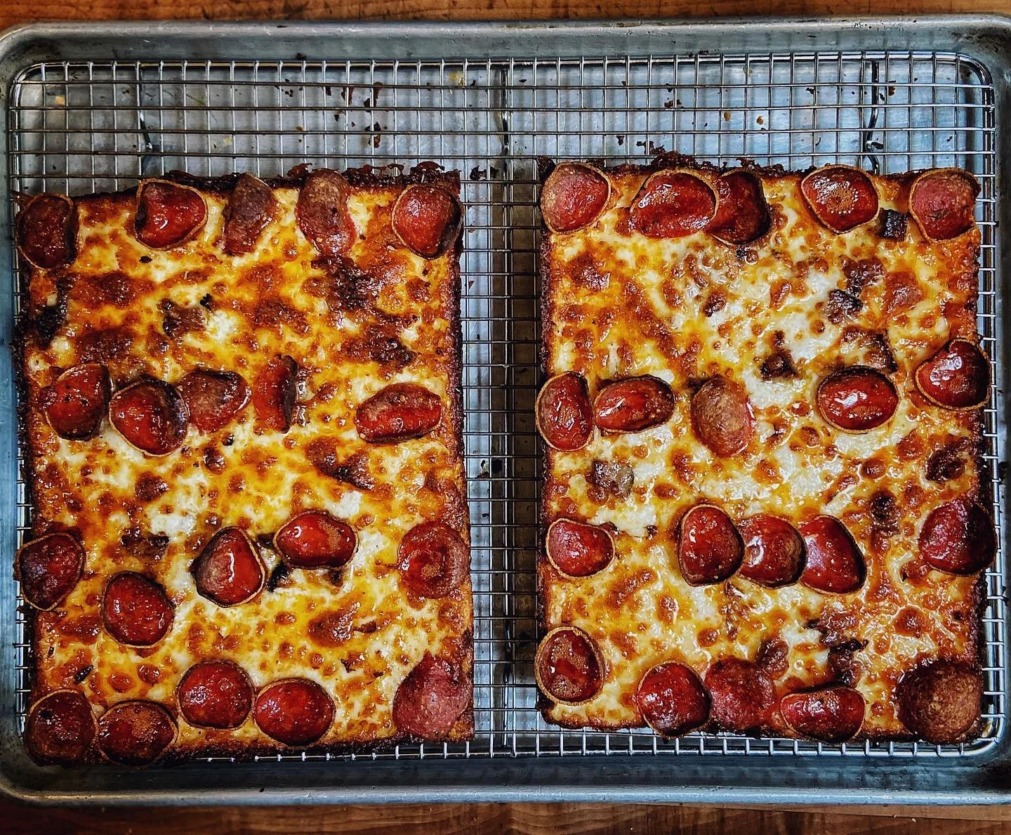 Two rectangular pepperoni and sausage pizzas on a wire cooling rack inside a metal baking sheet.